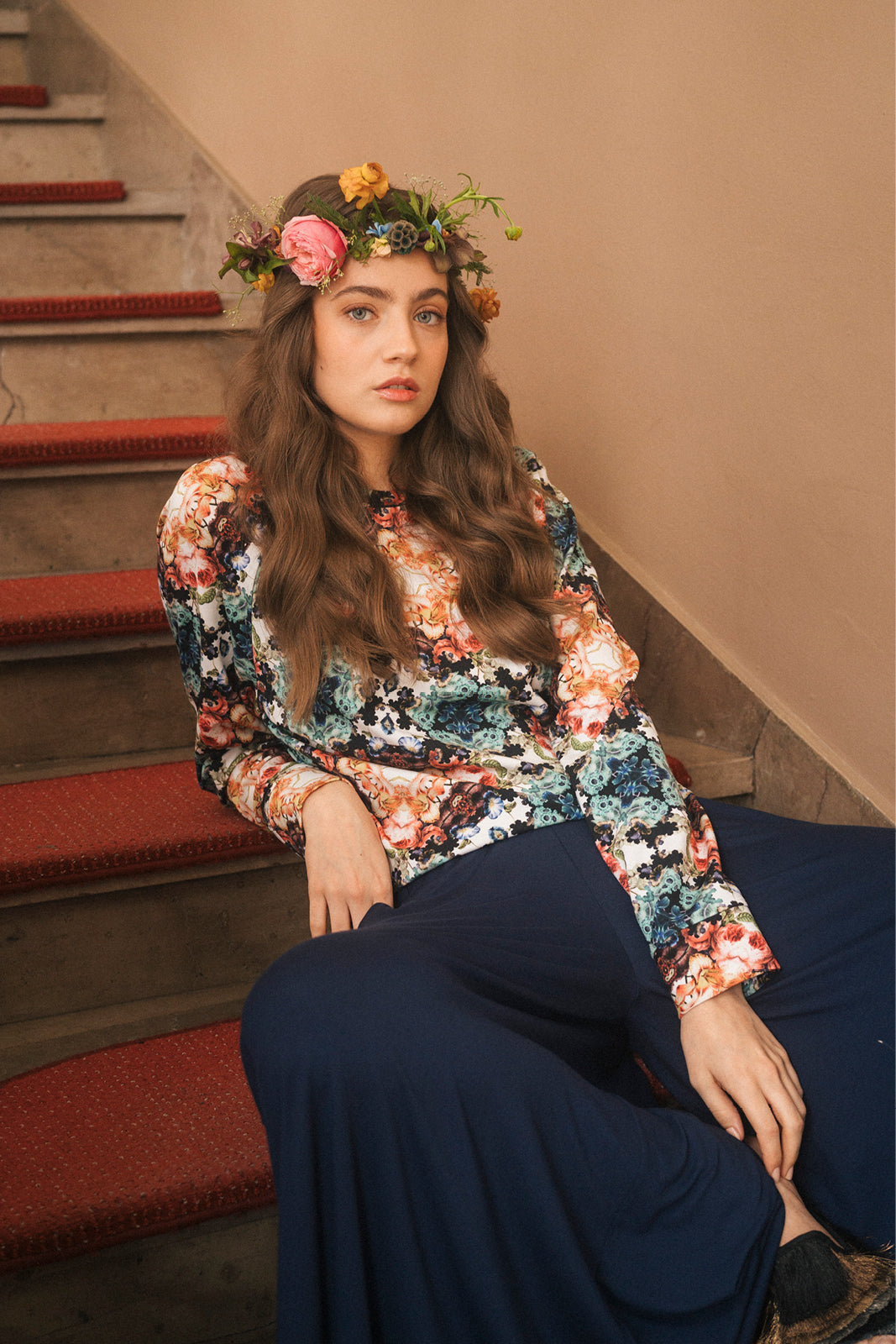 Kobieta w kwiecistej bluzce i granatowych spodniach, z wiankiem kwiatów na schodach – moda polska, self-care fashion